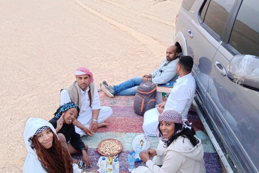 A group of people sitting on a carpeted area beside a vehicle, with a cooler and water bottles on the ground. 4