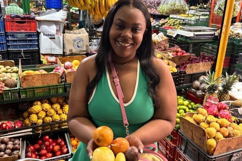 A person in a green top at a fruit market with various fruits and vegetables on display. 2