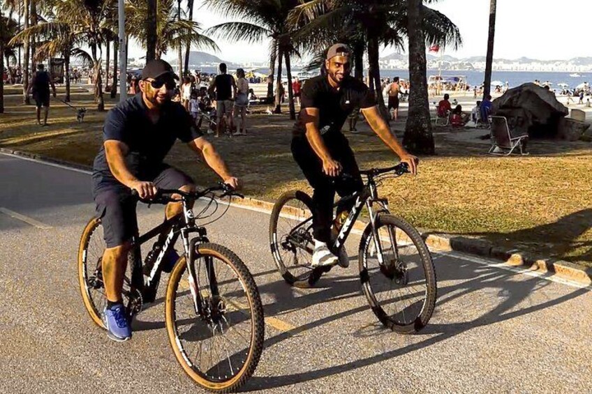 Two cyclists on a paved path with palm trees and a beach in the background. 5