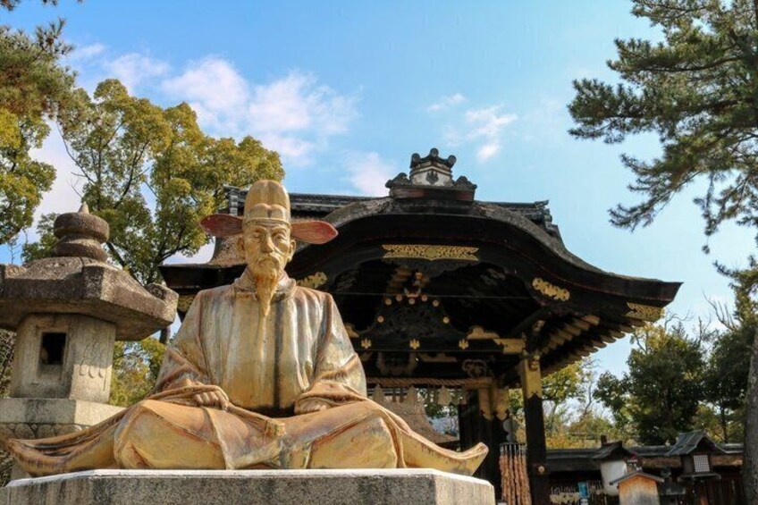 A statue of a seated figure with a traditional hat, a pagoda-style structure behind, and trees in the background. 1
