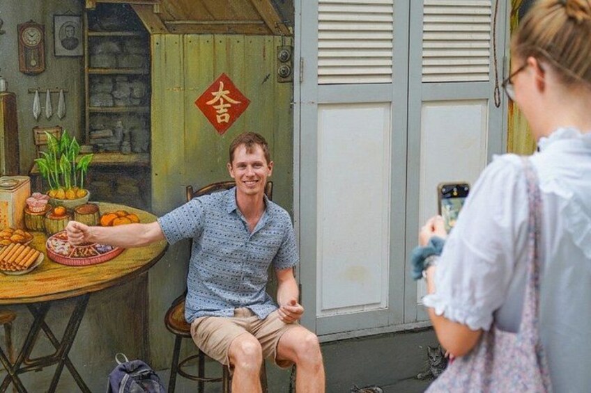 A man sitting at a table with food, a woman taking a photo, and a traditional Chinese symbol on the wall. 3