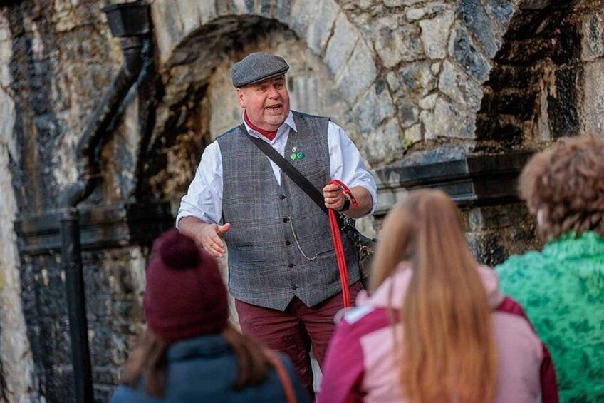 A man in a grey vest and cap speaking to a group of people. 3