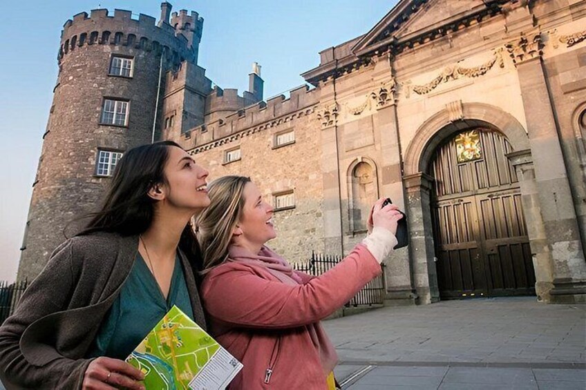 Two people taking a photo in front of a historic building with a large arched entrance and a tower. 1