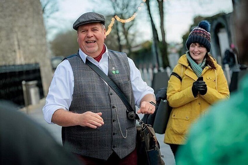 A man in a vest and hat, a woman in a yellow coat, and a street with buildings in the background. 2