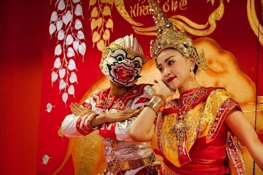 A person in traditional red and gold attire with a mask, standing in front of a red backdrop with gold designs. 1