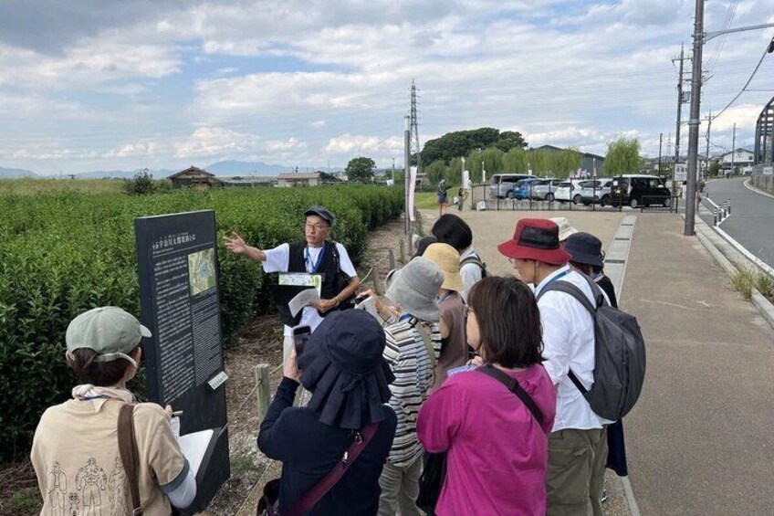 A group of people gathered around a signboard in a rural setting with fields and a road in the background. 1