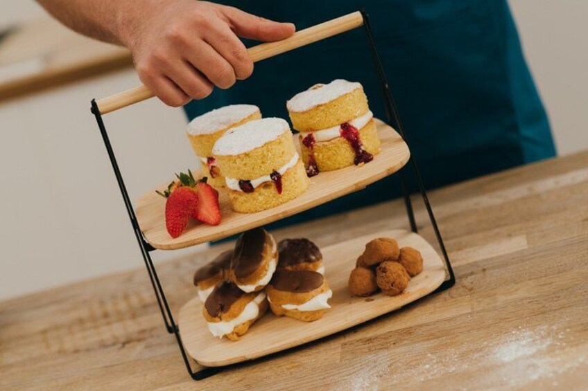 A wooden tiered stand with desserts, including sponge cakes with cream and strawberries, and chocolate-covered pastries. 1
