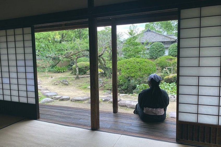 A person sitting by a traditional Japanese sliding door, overlooking a garden with rocks and greenery. 5