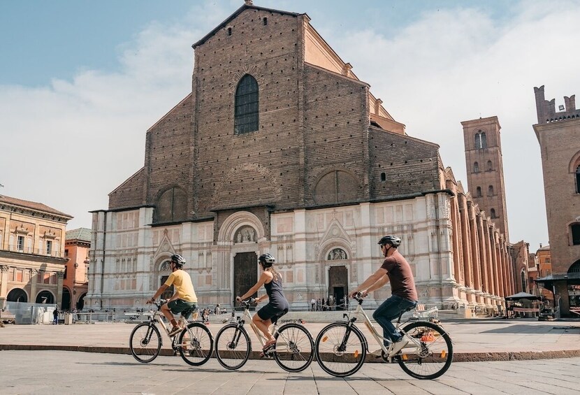 Three cyclists riding in front of a historic church with a bell tower. 1