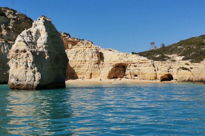 A coastal landscape with a prominent rock formation, a cave entrance, and a clear blue sea. 4