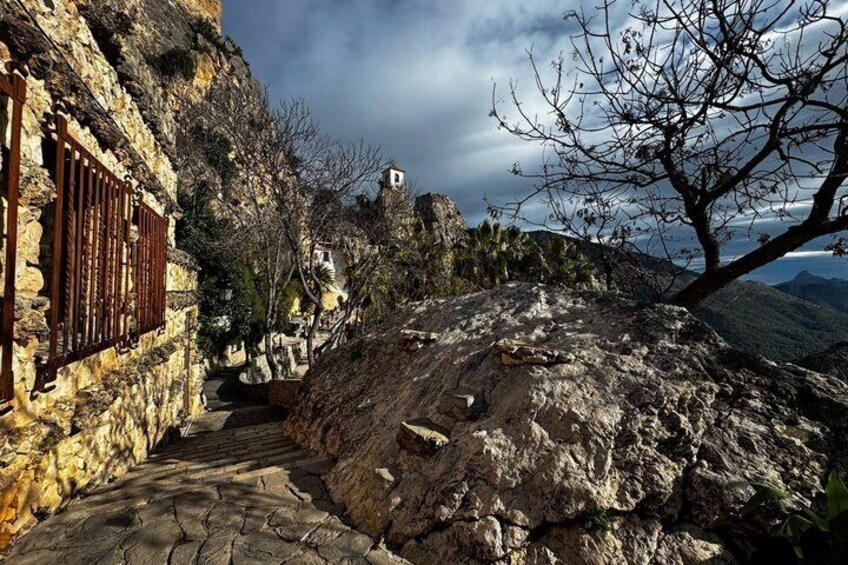 A stone building with a metal gate, a rocky path, and a tree with bare branches. 3