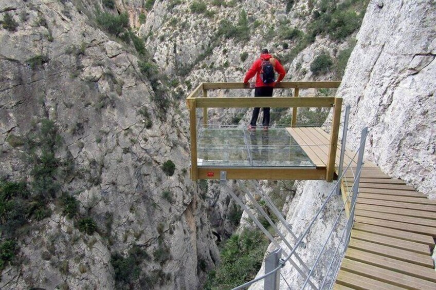 A person standing on a glass bridge with a wooden walkway. 1