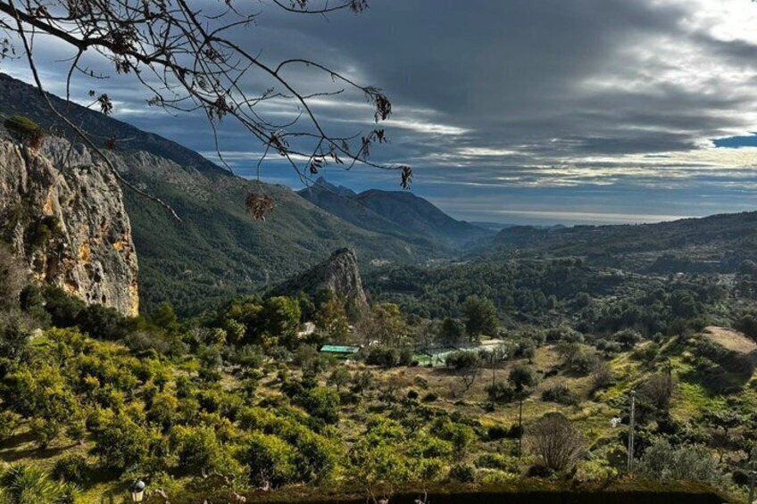 A mountainous landscape with a prominent rock formation, a valley with scattered trees, and a distant view of a house. 5