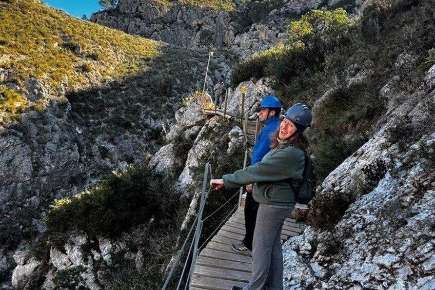 Two people on a wooden boardwalk in a rocky terrain with sparse vegetation. 2