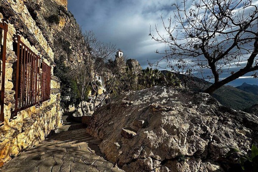A stone building with a wooden shutter, a rocky path, and a mountainous landscape in the background. 4