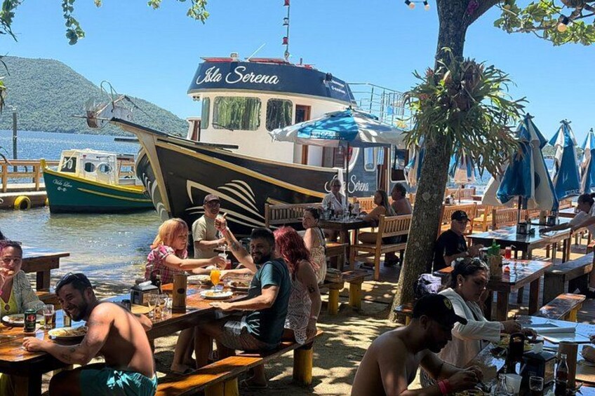 A waterfront dining area with people seated at tables, a boat named 'Isle Sereno' in the background, and a clear sky. 2