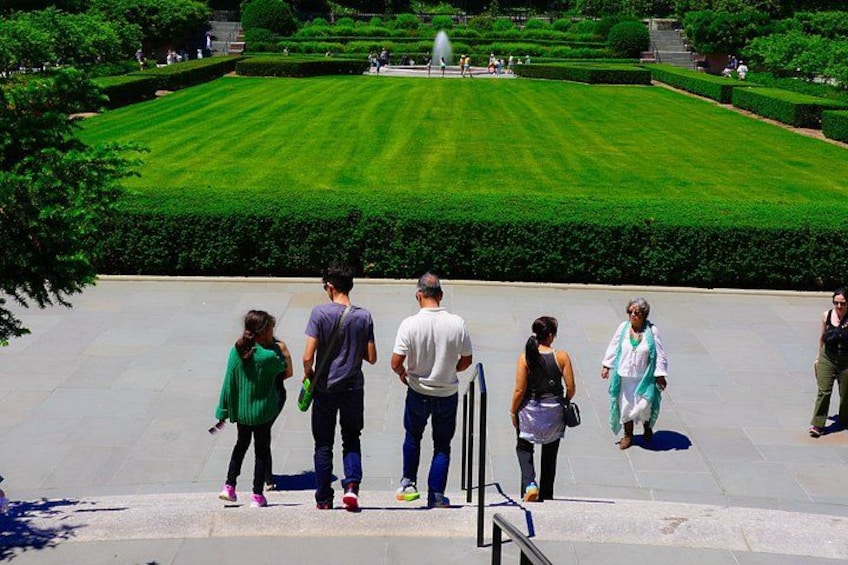 A group of people standing on a paved area overlooking a well-manicured lawn with a fountain in the background. 1