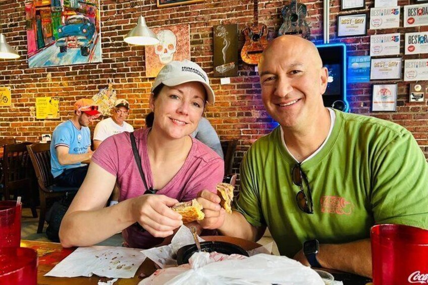 Two people sitting at a table with food, a brick wall, and various decorations in the background. 1