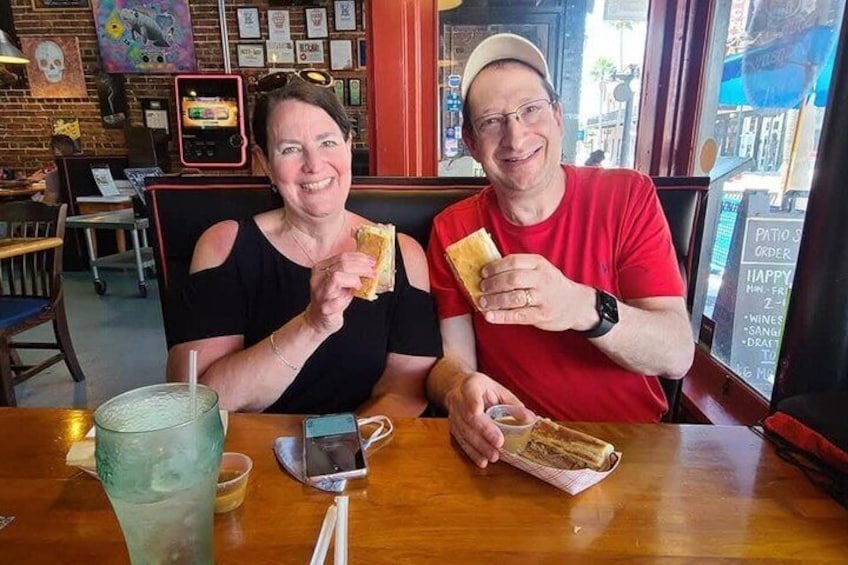 Two people sitting at a table with sandwiches, a drink, and a menu in a restaurant. 4