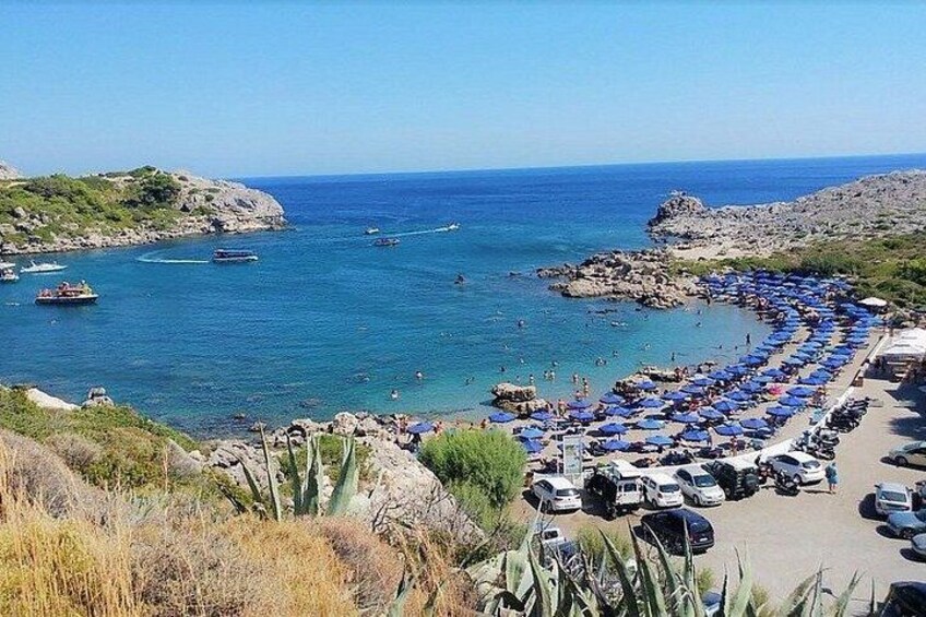 A coastal road with parked cars and blue umbrellas, leading to a beach with boats in the water. 3