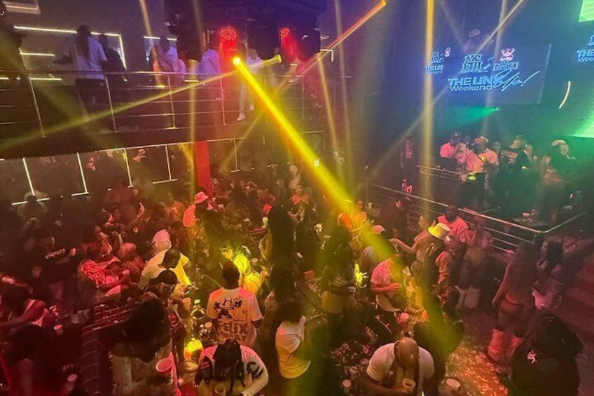 A crowded indoor event with a stage and a DJ booth. 3