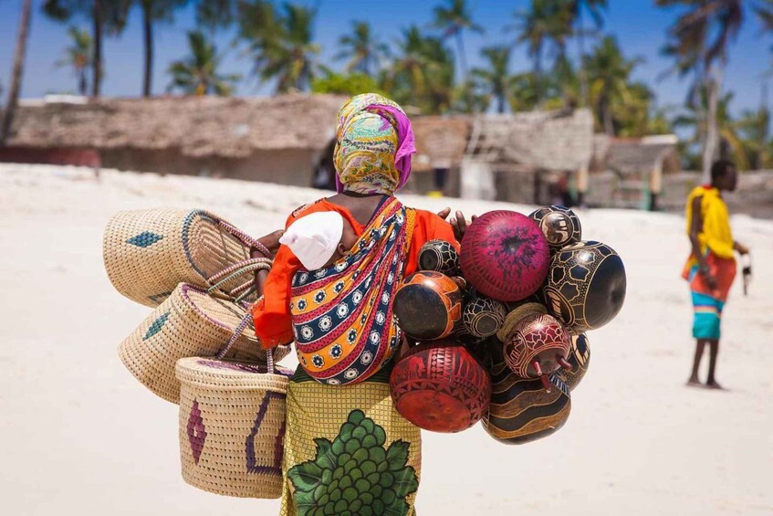 A person carrying a variety of woven baskets and pottery on a sandy beach with palm trees in the background. 5