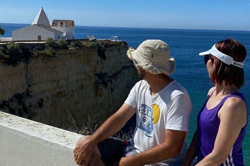 Two people sitting by a cliff overlooking the ocean, with a white building and clear blue sky in the background. 5