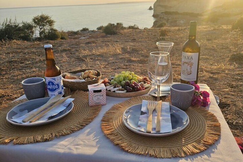 A picnic setup with plates, cutlery, a bottle, and glasses on a woven mat by the sea. 2
