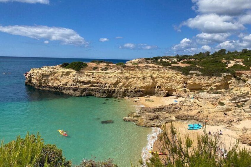 A coastal landscape with rocky cliffs, clear turquoise waters, and a sandy beach with a blue umbrella. 1
