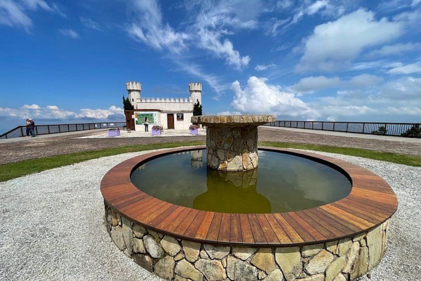 A circular wooden deck with a stone fountain in the center, surrounded by a stone wall, with a castle-like building in the background. 4