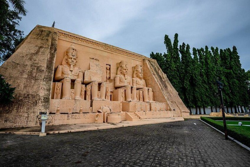 A large stone structure with seated figures, surrounded by trees and a paved area. 3