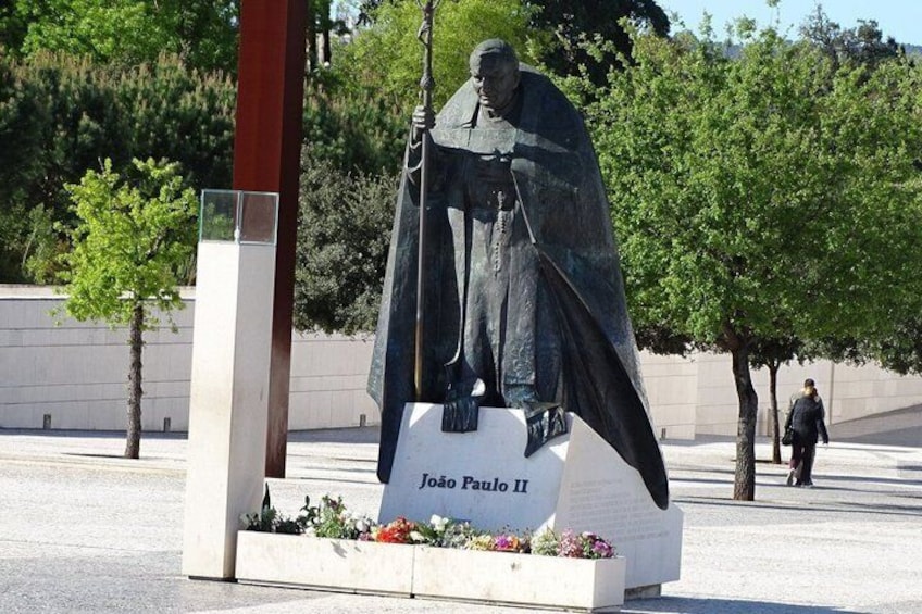 A statue of João Paulo II in a public square with trees and a white building in the background. 4