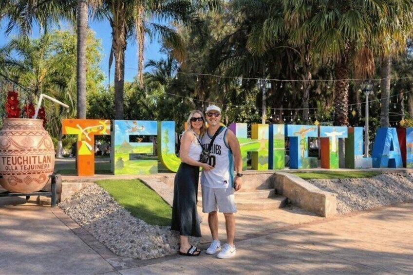 A couple standing in front of colorful letters spelling 'TEUCHITLAN' with palm trees and a decorative pot in the background. 1