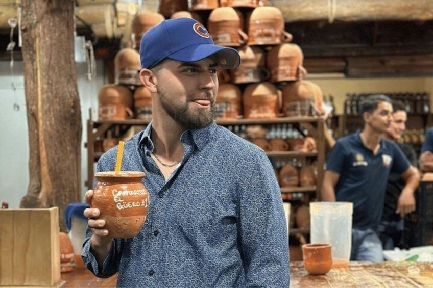 A person holding a clay pot with a straw, in a market with pottery items in the background. 5