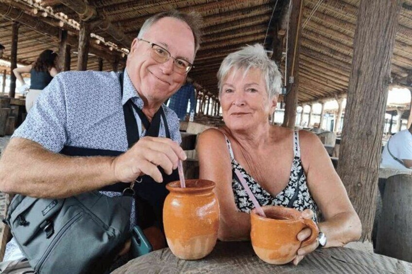 Two people sitting at a table with a large clay pot and a straw. 2