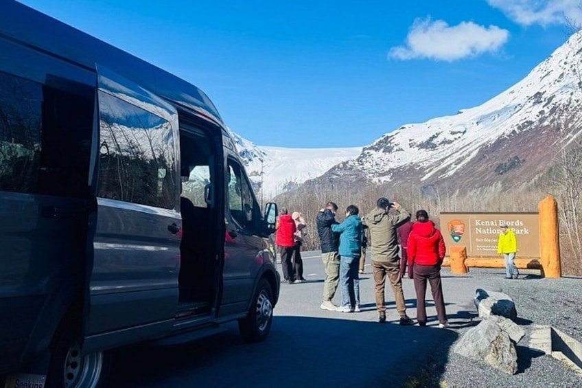 A group of people standing near a black van and a sign for Kenai Fjords National Park. 4