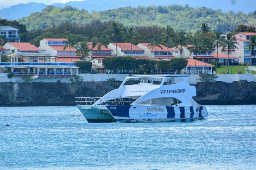 A boat named 'North Bay' with 'VIP EXPERIENCE' written on it, floating on water with a coastal town and palm trees in the background. 1