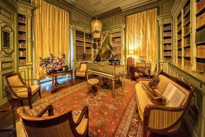 A room with a grand piano, a sofa, chairs, a table with flowers, and bookshelves filled with books. 5