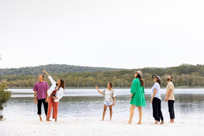 A group of people standing on a sandy beach by a lake with trees in the background. 1