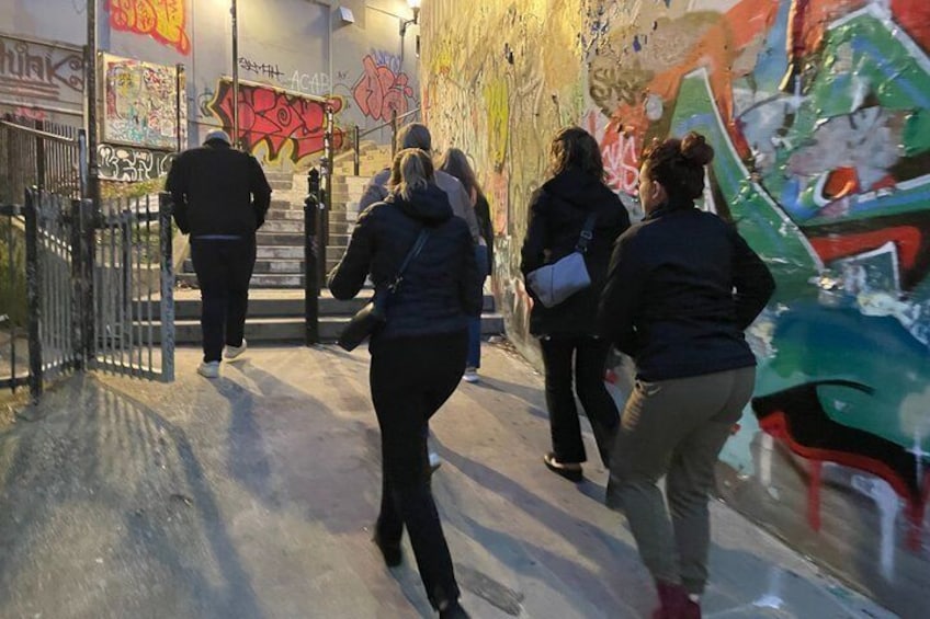 A group of people walking on a street with graffiti-covered walls and a metal fence. 2