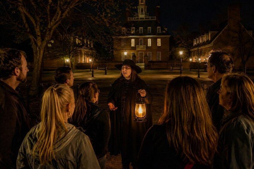 A group of people gathered around a person holding a lantern in a historic town square at night. 1