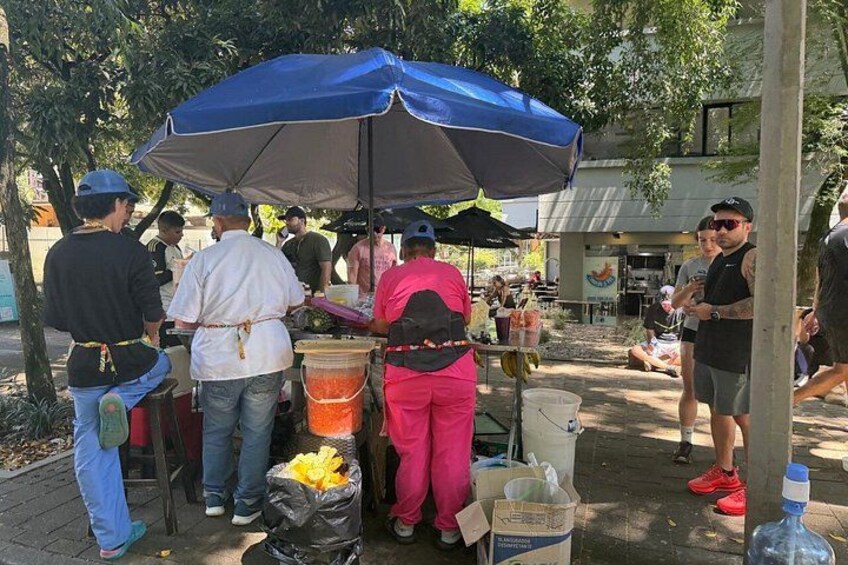 A street food vendor setup with a blue umbrella, food containers, and a crowd of people. 2