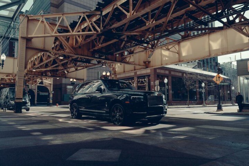 A black luxury car parked on a city street under a large metal bridge. 3