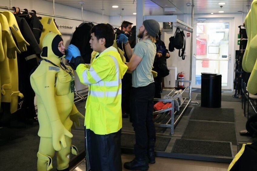 A person in a yellow hazmat suit, another in a yellow jacket, and a third in a grey hoodie in a room with yellow suits on racks. 3