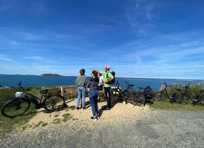 Three people standing by a coastal road with bicycles parked nearby, overlooking the sea. 1