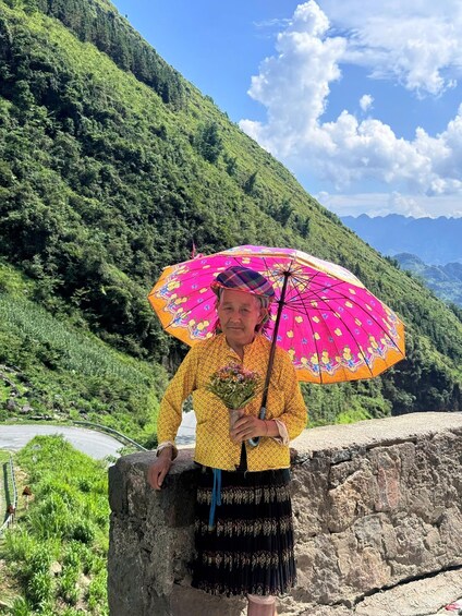 A person holding a colorful umbrella, standing on a stone ledge with a mountainous landscape in the background. 2