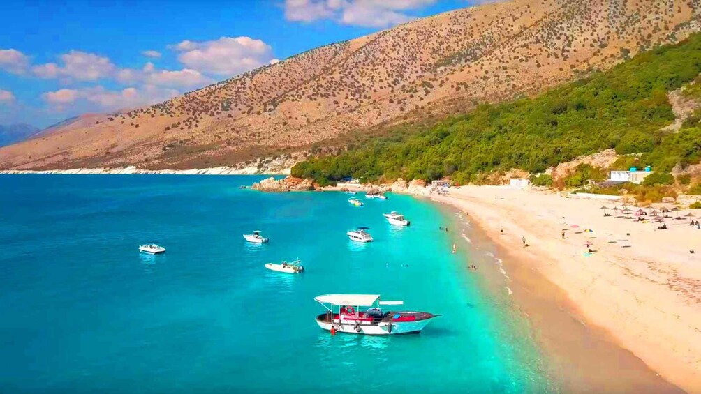 A coastal beach with boats anchored near the shore, a sandy beach with umbrellas, and a mountainous backdrop. 1