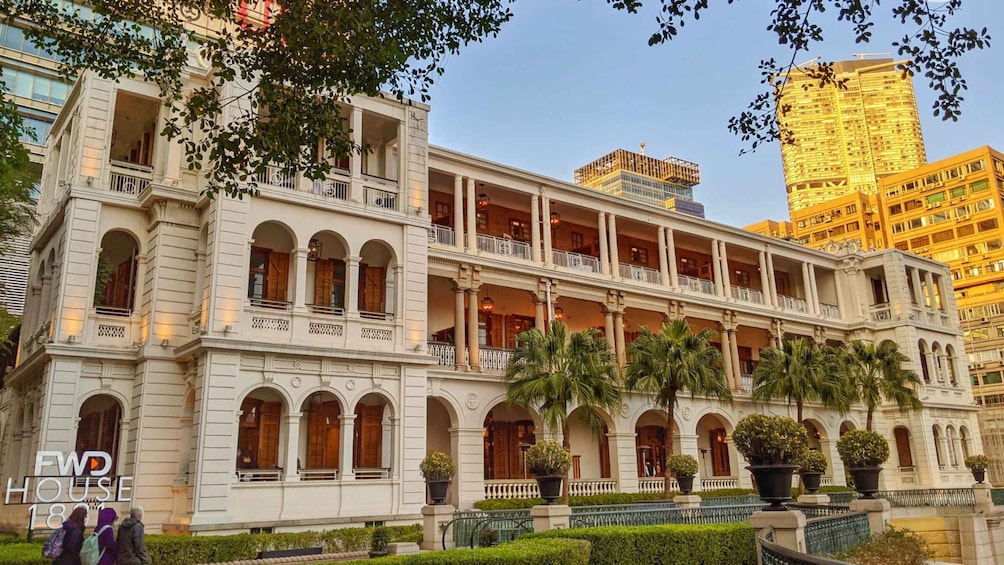 A historic building with multiple balconies and columns, surrounded by palm trees and modern high-rise buildings. 1