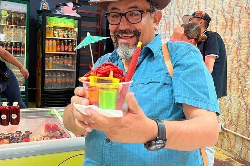 A person holding a colorful drink with a straw, a beverage dispenser in the background, and a fence. 2