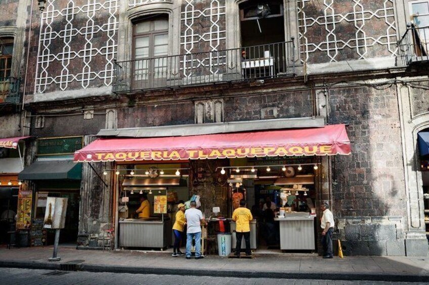 A street-side taco stand with a red awning and a few customers. 2
